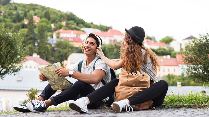 Young man holding a map, wearing a backpack and headphones, sitting on a cobblestone pathway next to a young woman playfully trying to remove his headphones, with a few hillside country homes in the background.