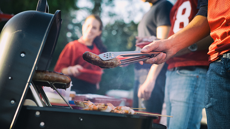 Cuatro personas cocinando comida en una parrilla al aire libre, una de ellas sosteniendo una salchicha sobre la parrilla con un par de pinzas.