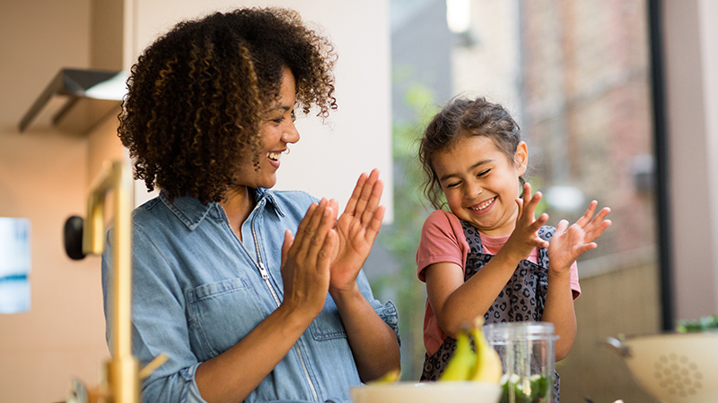 Mamá, papá e hija sonrientes en una cocina rústica frente a una barra con un bol de ensalada, una tabla de picar y otras verduras y condimentos.