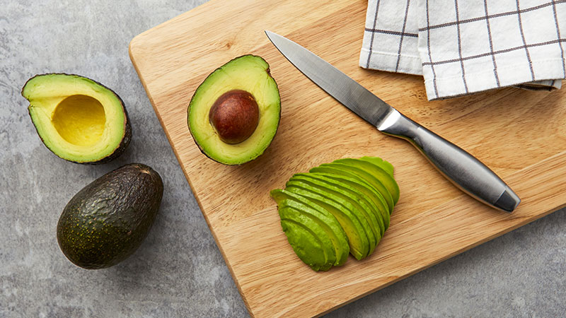 A whole avocado, two avocado halves, and avocado slices on a wooden chopping board next to a knife.