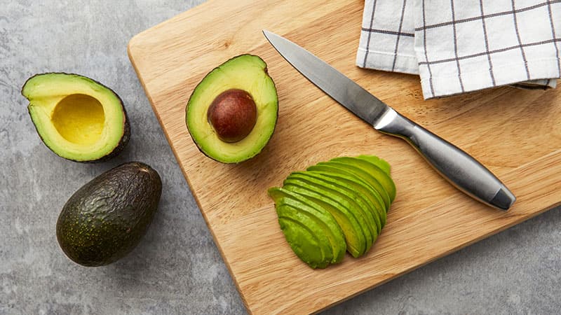 Un aguacate entero, dos mitades de aguacate y rebanadas de aguacate sobre una tabla de picar, junto a un cuchillo.