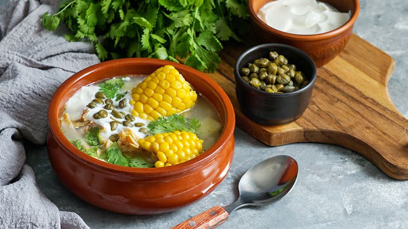 Colombian Ajiaco soup with ears or corn served on a clay bowl, next to a spoon and a wooden board with spices and fresh herbs.